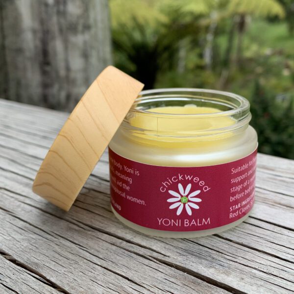 Image showing a glass jar filled with pale yellow yoni balm with the lip propped on the side. The jar has a red label, and is on a wooden outdoor table, with garden in the background.